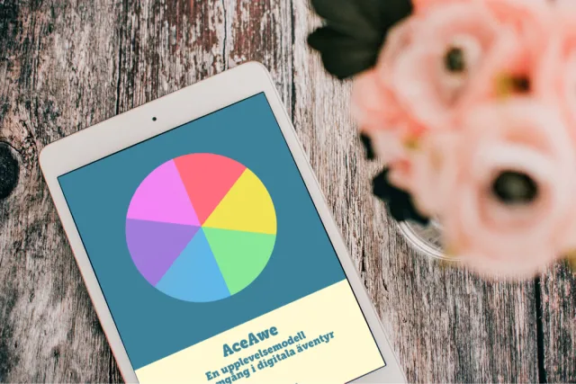 E-boken AceAwe – en upplevelsemodell för framgång i digitala äventyr, på en surfplatta på ett träbord bredvid rosa blommor. Omslaget visar en cirkel i sex färger.