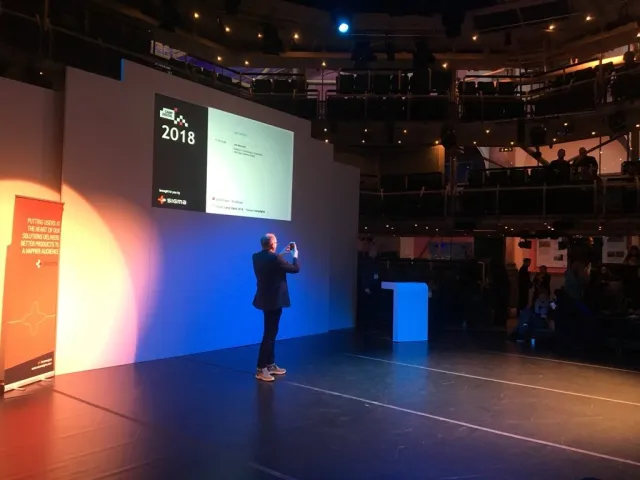 Photo: Katy Arnold: Per Axbom on stage taking a photo of the audience, with the Camp Digital 2018 slide behind him.