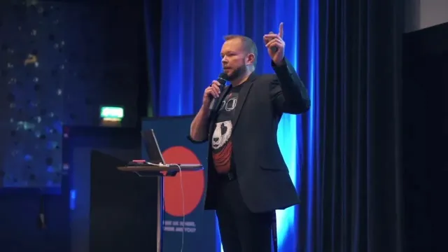 Per Axbom on a dark stage, pointing upwards with a microphone in his other hand.