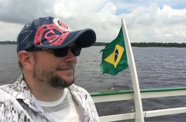 Close-up of Per Axbom in a baseball cap and sunglasses on a boat, with water behind him and land in the distance.