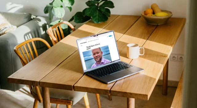 A laptop on a sunlit wooden kitchen table, next to a coffee mug and a bowl of fruit. On the screen: Svenska Dagbladet's debate section with the headline “Reglering av AI är ett måste” and a photo of Per.