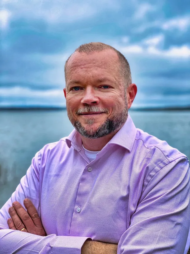 Photo: Simon Axbom: The portrait published with the article: Per in a lilac shirt, arms crossed, smiling, by a lake under a cloudy blue sky.