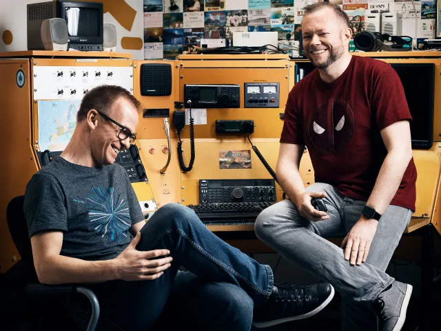 Per Axbom (right, in a red t-shirt) and James Royal-Lawson laughing at an orange radio desk full of amateur-radio equipment.