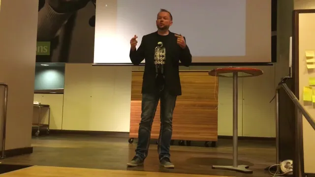 Still from the recording: Per on stage in jeans, a dark jacket and a T-shirt, gesturing with both hands, with an empty projector screen behind him – the talk was given without slides.